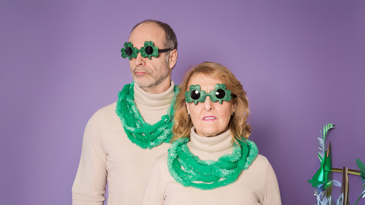 A couple wearing matching beige turtlenecks, green scarves, and festive shamrock-shaped glasses, posed against a purple background