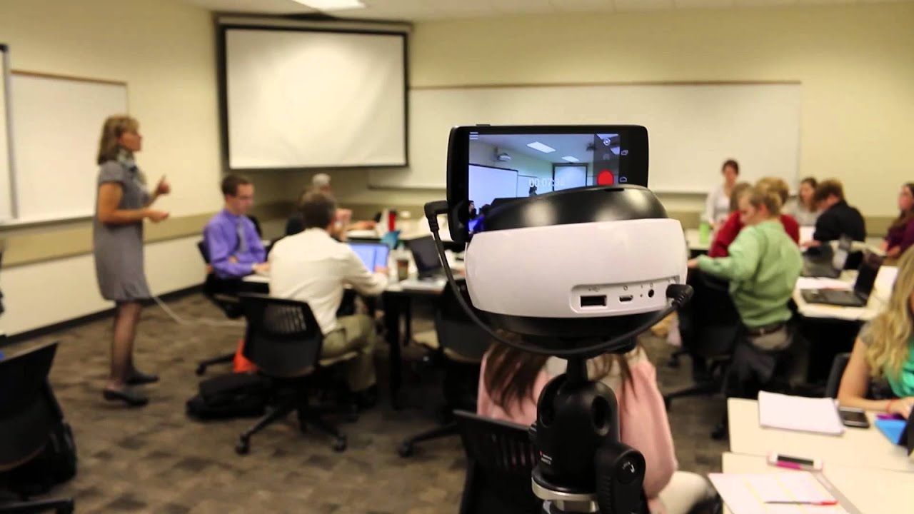 A Swivl robot on a tripod with a smartphone mounted on top, tracking a teacher in a classroom while students sit at tables.